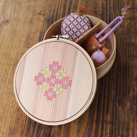 A wooden box with a round lid, containing various small items such as a pink and white flower-patterned pouch, a small pink ball, and a small brown leather object.