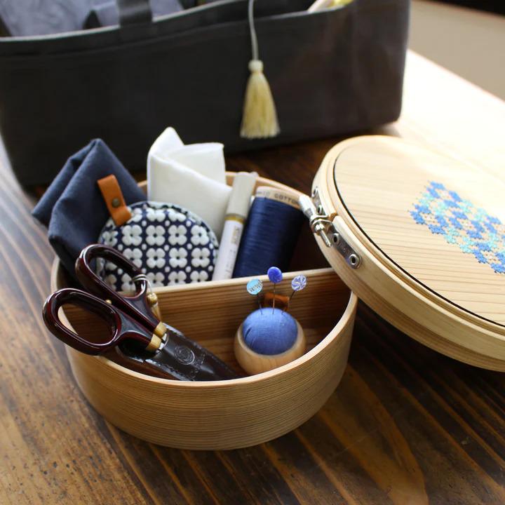 A wooden container filled with various sewing and crafting supplies, including a pair of scissors, a needle, thread, pins, and a tassel.