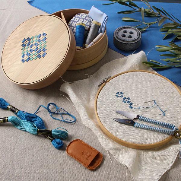 various sewing and crafting supplies, including a wooden box with embroidery floss, a wooden hoop with a needle and thread, and a pair of scissors.