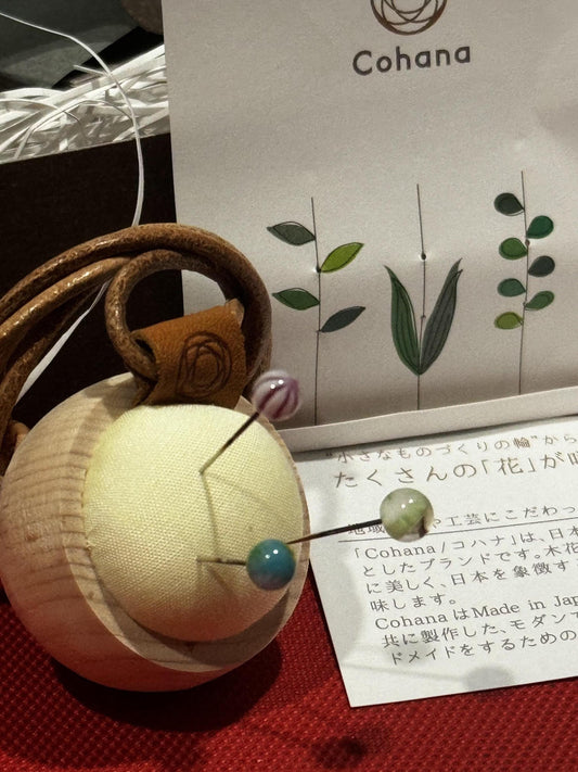 a small, round, white object with a brown leather strap, a needle, and two blue and green beads, all placed on a red surface. There is also a piece of paper with Japanese text and a plant illustration in the background.