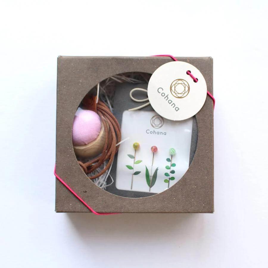 A brown cardboard box with a pink and white label, containing a small pink ball, a white card with a floral design, and a small green plant.