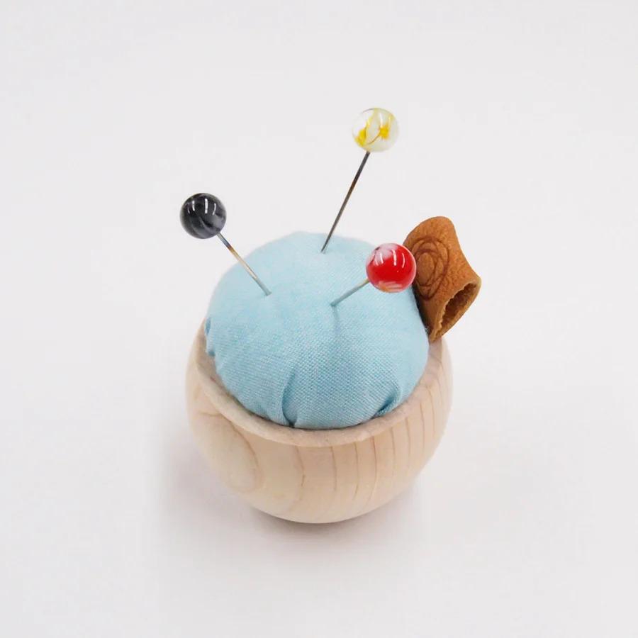 A wooden pin cushion with three colorful pins inserted into it, sitting on a white surface.