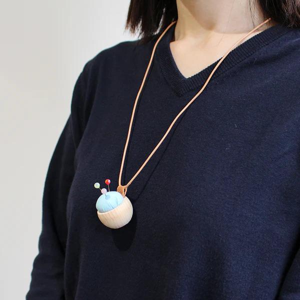 A person wearing a black sweater with a wooden pincushion pendant necklace.