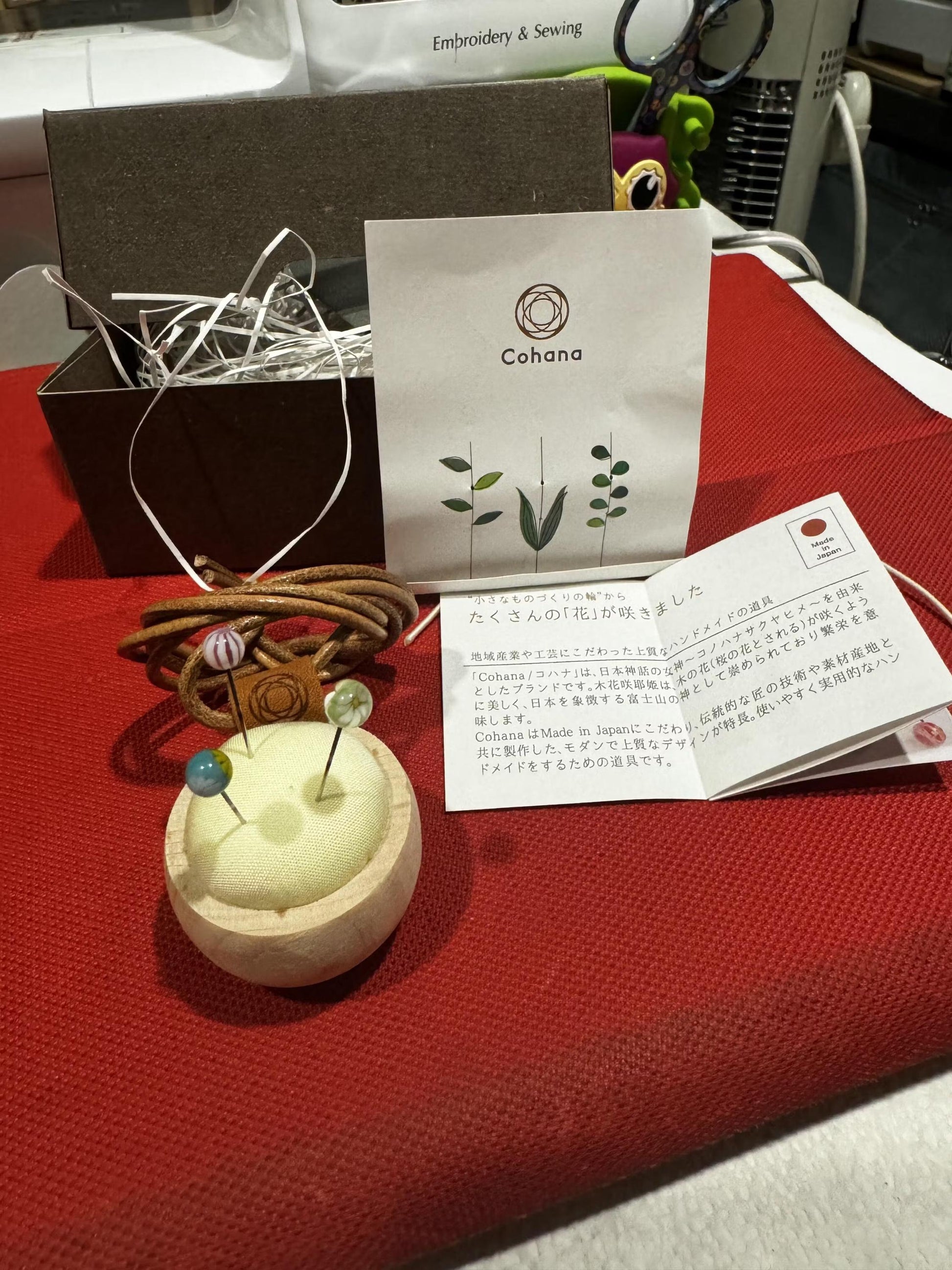 a craft kit on a red tablecloth, including a wooden pin cushion, a spool of thread, and a card with text and illustrations.