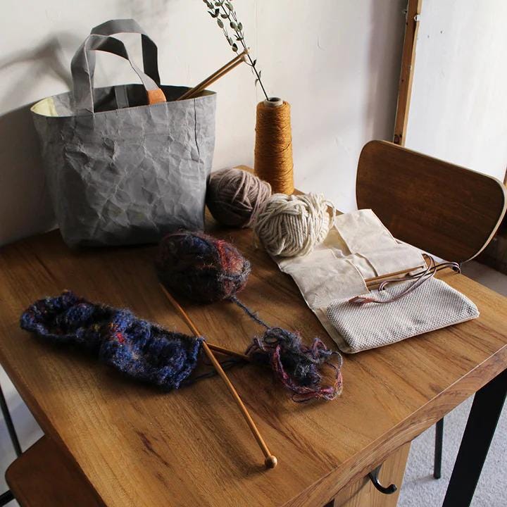 A wooden table with various crafting supplies, including yarn, knitting needles, and a gray tote bag, suggesting a creative workspace.