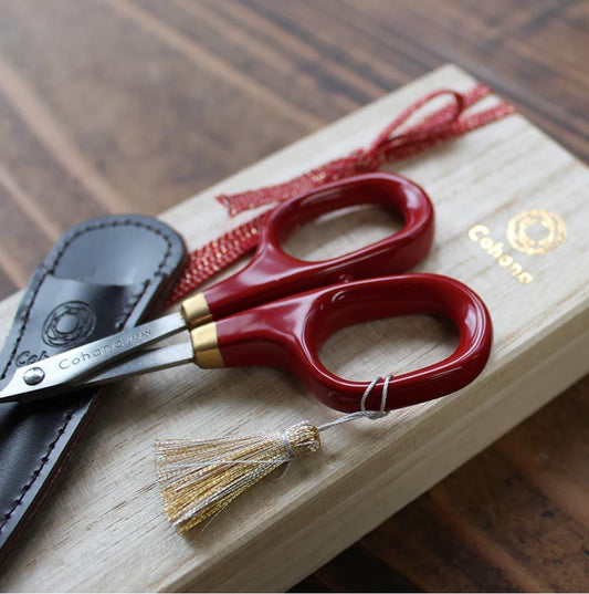A pair of red-handled scissors with a tassel and a black leather case with a gold-colored logo are placed on a wooden surface.
