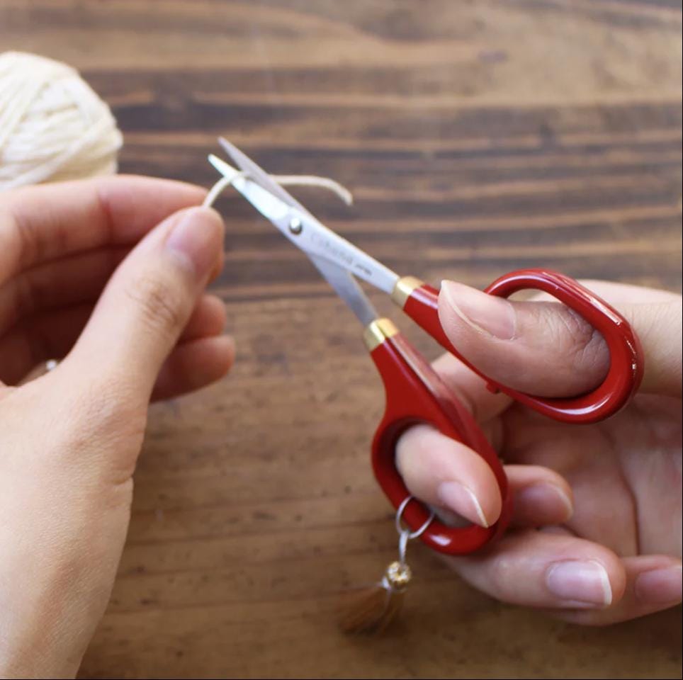 A person's hands holding red scissors and cutting a string of yarn on a wooden surface.