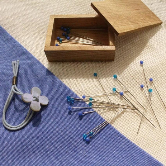a wooden box filled with blue pins, a white flower-shaped pin, and scattered blue pins on a blue and white striped fabric.