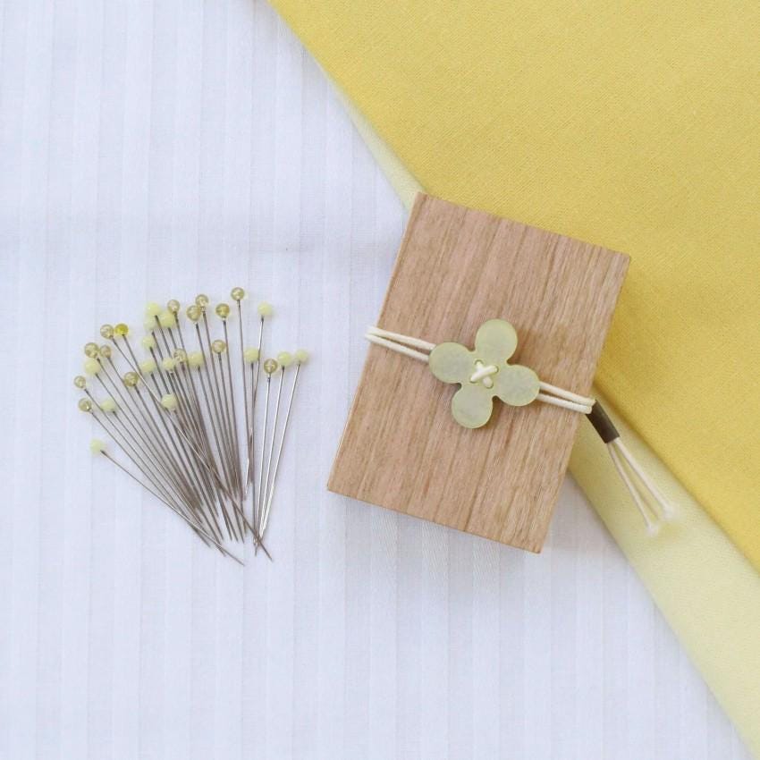 a collection of small, yellow pins and a wooden box with a flower-shaped button on it, all placed on a white striped fabric background.