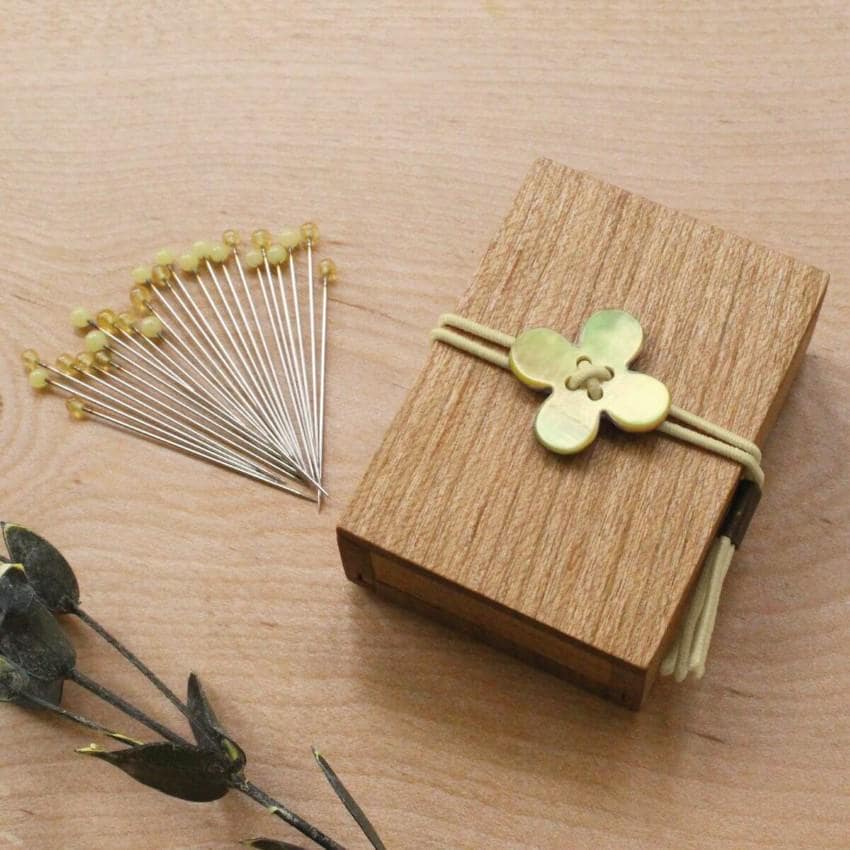 a wooden box with a flower-shaped button on it, surrounded by a collection of yellow pins and a sprig of green leaves on a wooden surface.