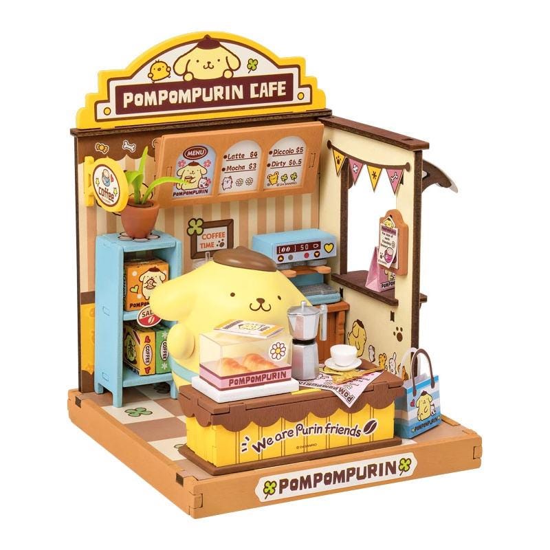 A small, colorful toy cafe featuring a yellow pom-pom character serving coffee and pastries, with various other small items and decorations.