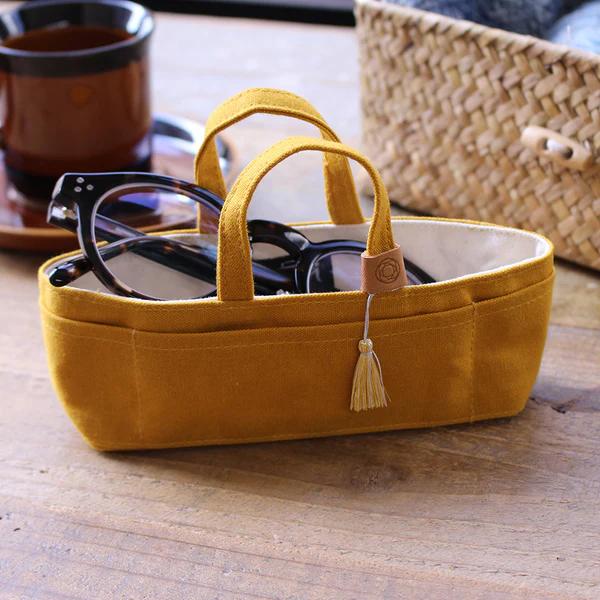 A yellow tote bag with a tassel hanging from it, containing a pair of glasses, placed on a wooden surface.