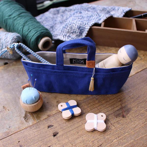 A blue tote bag with a tassel, a blue and white striped pin cushion, and wooden buttons are arranged on a wooden surface.