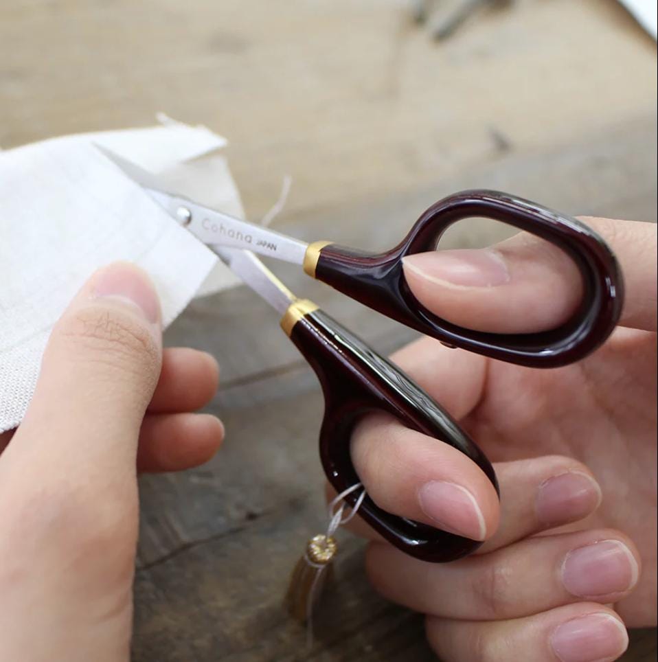 A person's hand holding a pair of red-handled scissors with gold accents, cutting a piece of white fabric.