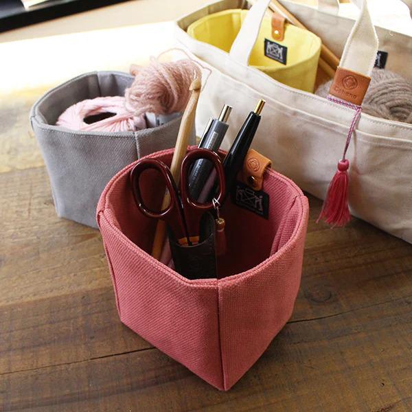 A pink fabric container holding various craft supplies, including scissors, pens, and yarn, sits on a wooden surface.