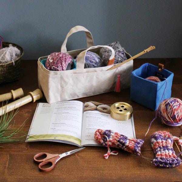 A beige tote bag filled with yarn and knitting needles sits on a wooden surface, accompanied by a book, scissors, and a small blue container.