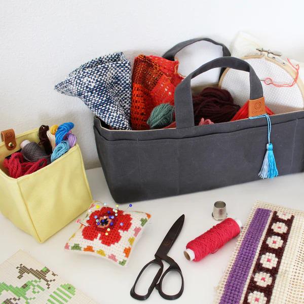 a variety of craft supplies, including a gray tote bag, a yellow container, a pair of scissors, a cross-stitch kit, and a sewing kit, all arranged on a white surface.