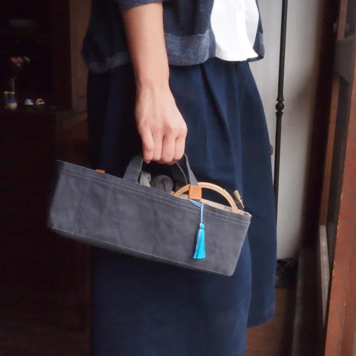 A person is holding a gray tote bag with a blue tassel attached, standing in front of a wooden door.