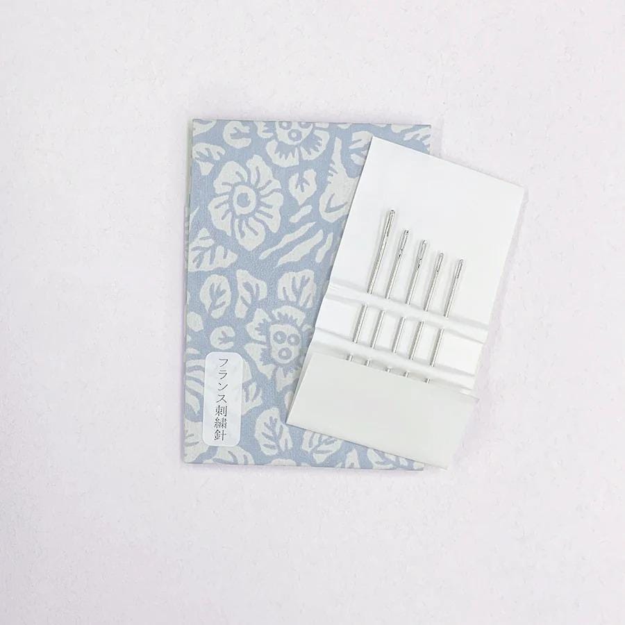 A blue and white patterned notebook and a white sewing kit with four needles are shown against a white background.