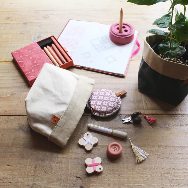 a variety of craft supplies and personal items arranged on a wooden surface, including a pink box, a notebook, a potted plant, a hat, a pencil, a small purse, a tassel, a button, and a pair of scissors.
