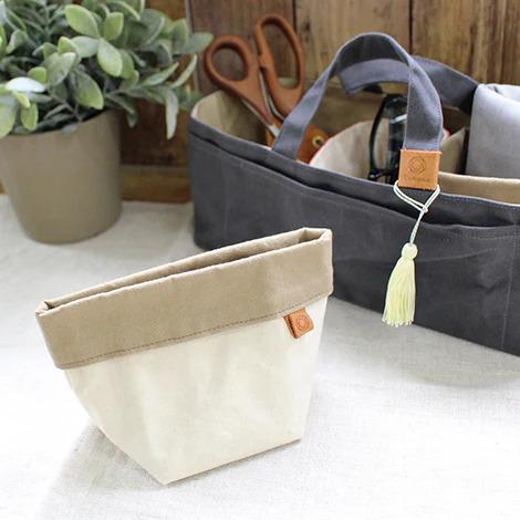 a beige and gray canvas tote bag with a tassel hanging from it, placed next to a pair of scissors and a potted plant.