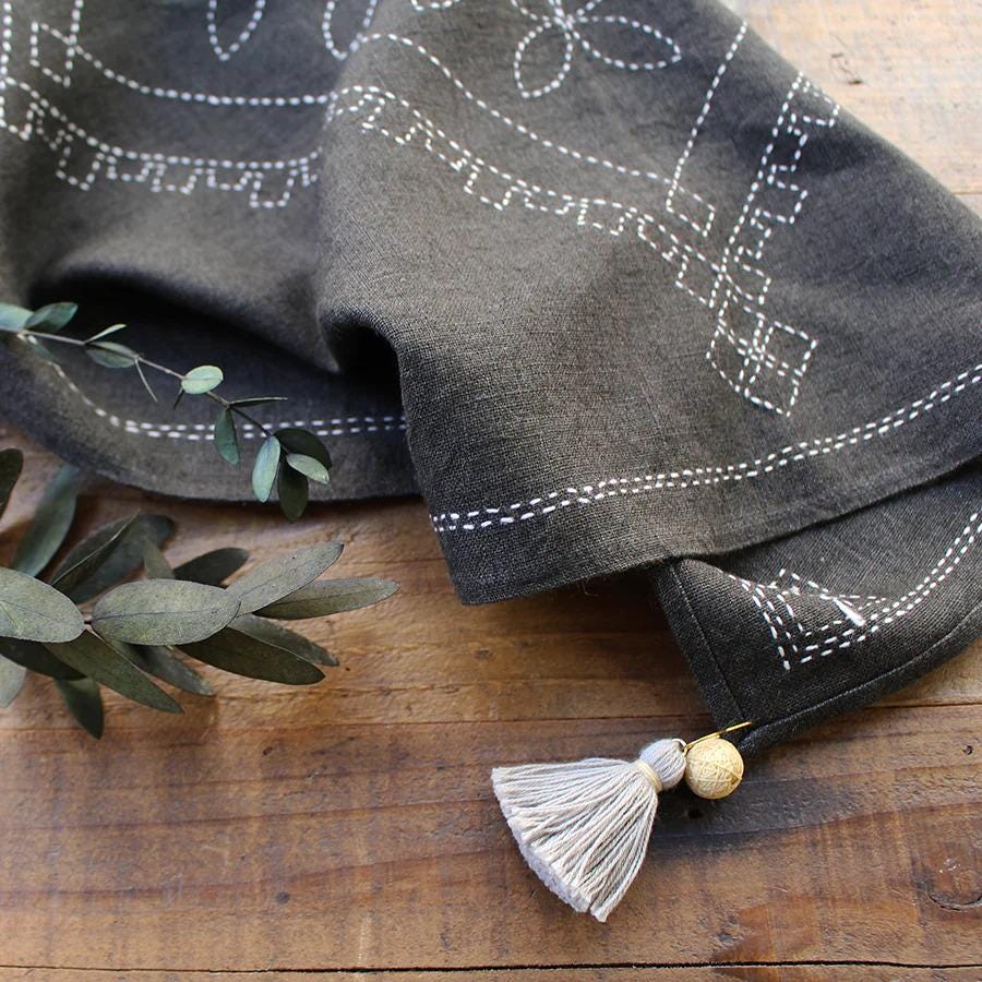 a black fabric with white embroidered designs, a white tassel, and some greenery on a wooden surface.