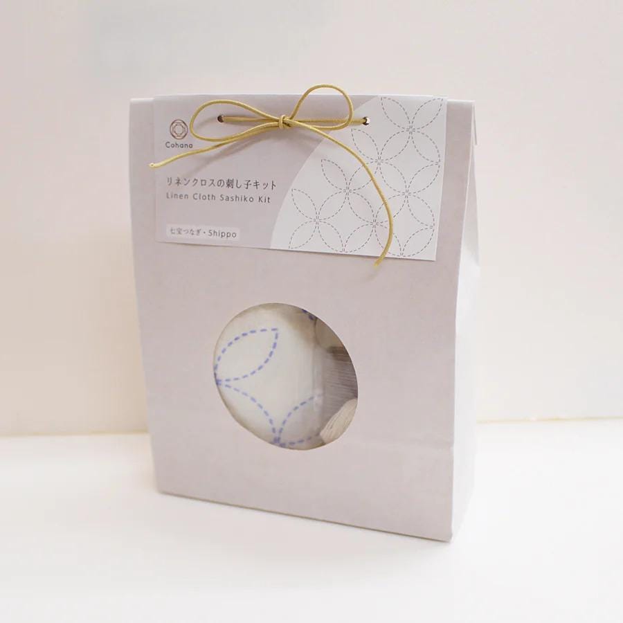 A white paper package with a gold bow, containing a white cloth with blue stitching and text in Japanese.