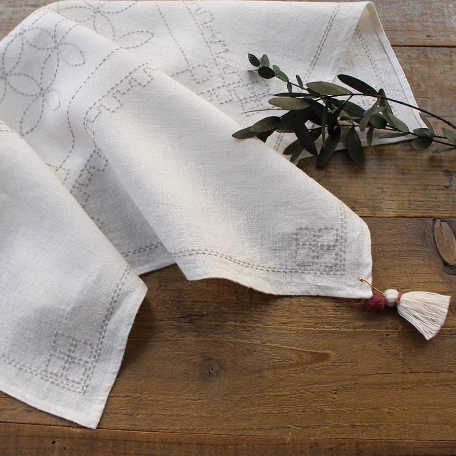 a white handkerchief with a tassel hanging from it, placed on a wooden surface alongside a branch of greenery.