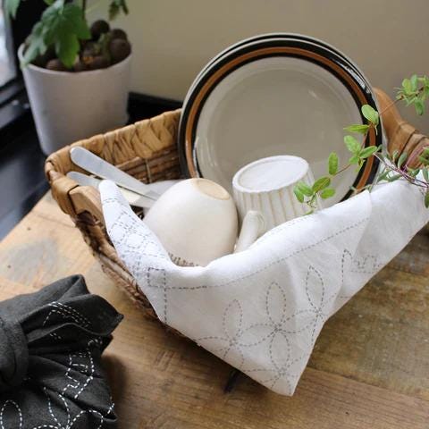 A wicker basket filled with various kitchen items, including plates, a teapot, and a plant, sits on a wooden surface.