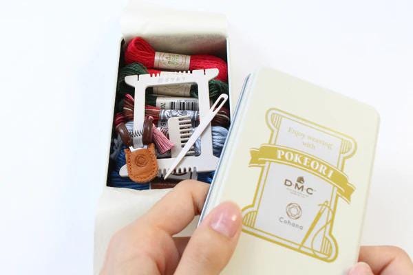 A hand holding a box of Pokemon cards, with a spool of thread and a needle visible inside the box.