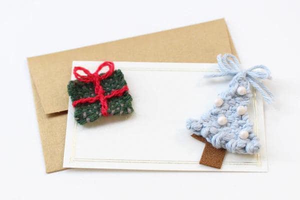 a white card with a green and red knitted gift box and a blue knitted Christmas tree ornament placed on top of it.