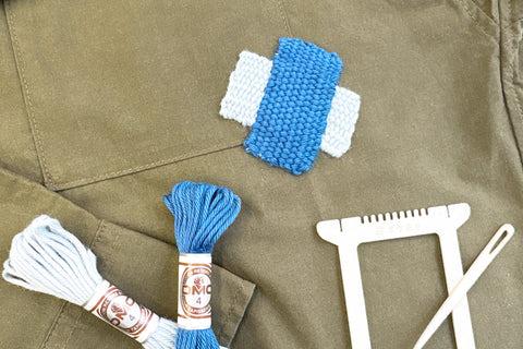 a variety of crafting supplies, including a spool of blue thread, a spool of white thread, a needle, and a frame, all arranged on a brown fabric surface.