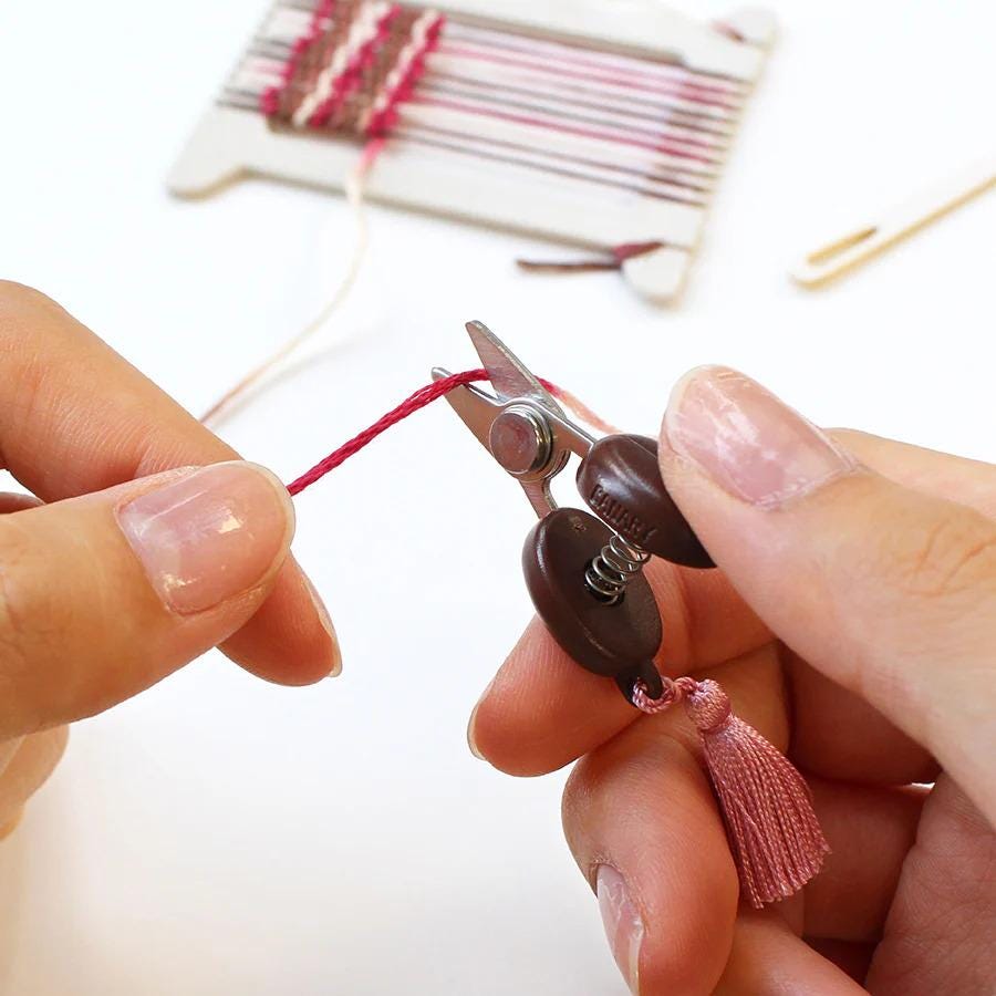 A person's hands are carefully cutting a red string with a pair of scissors, with a tassel attached to the end of the string.