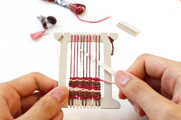 A person's hands are holding a small weaving frame with red and brown threads, and they are weaving a small piece of fabric on the frame.