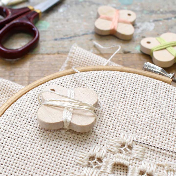 a close-up view of a needle and thread, with a wooden butterfly-shaped pin and a cross-stitch pattern on a white fabric.