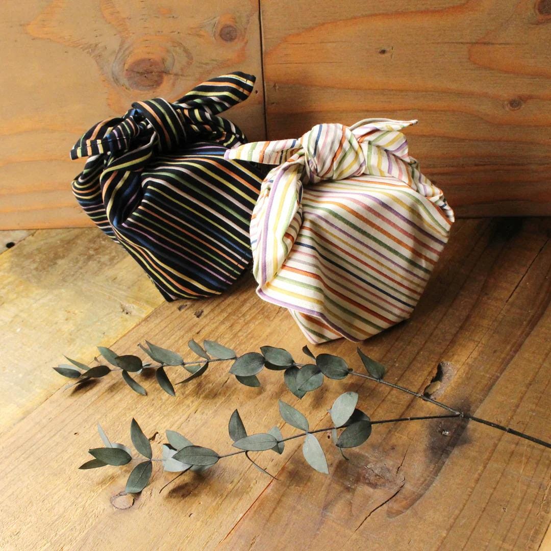 Two wrapped gifts with colorful striped fabric are placed on a wooden surface, accompanied by a sprig of greenery.