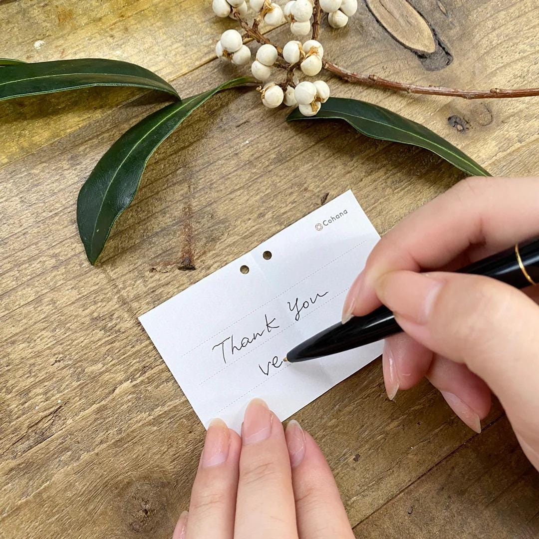 A person's hand is seen writing a handwritten note on a piece of paper, with a pen in use, on a wooden surface.