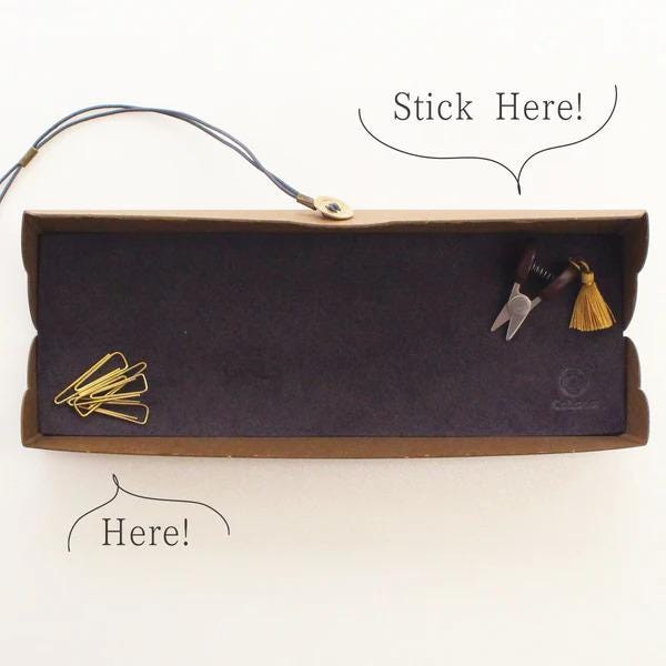 a brown cardboard box with a black interior, containing various office supplies such as a stapler, scissors, and a tassel. There are also two speech bubbles above the box, one saying "Stick Here!" and the other saying "Here!".