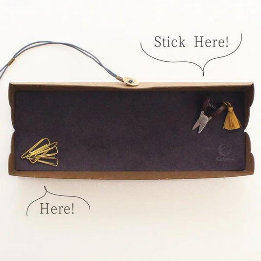 a brown cardboard box with a black interior, containing various office supplies such as a stapler, scissors, and a tassel. There are also two speech bubbles above the box, one saying "Stick Here!" and the other saying "Here!".