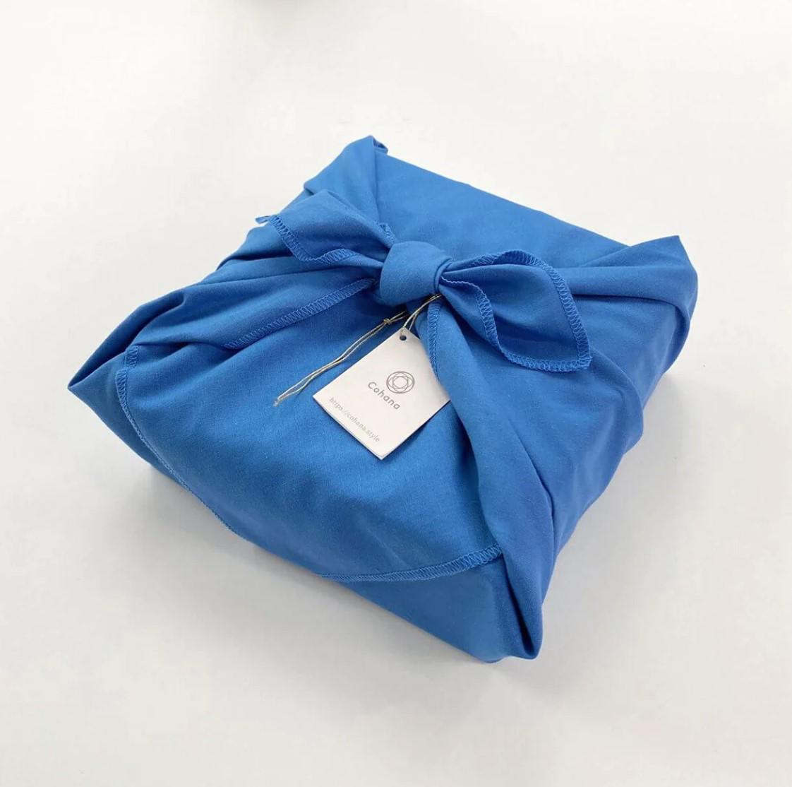 A blue gift box wrapped in a blue cloth with a white tag attached, placed on a white surface.