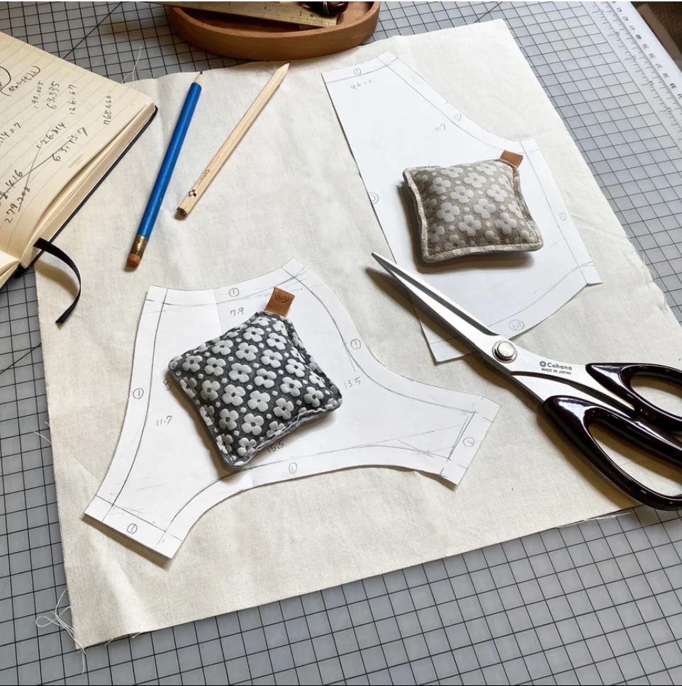 a sewing or crafting workspace with various tools and materials, including a pattern, a small fabric piece, a pair of scissors, and a notebook.