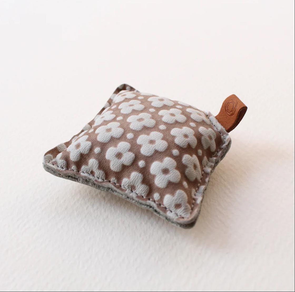 A small, square-shaped object with a brown floral pattern and a brown leather tag attached to it, resting on a white surface.