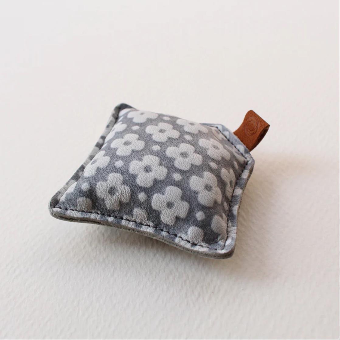 A gray and white floral patterned fabric item with a brown leather tag attached to it, resting on a white surface.