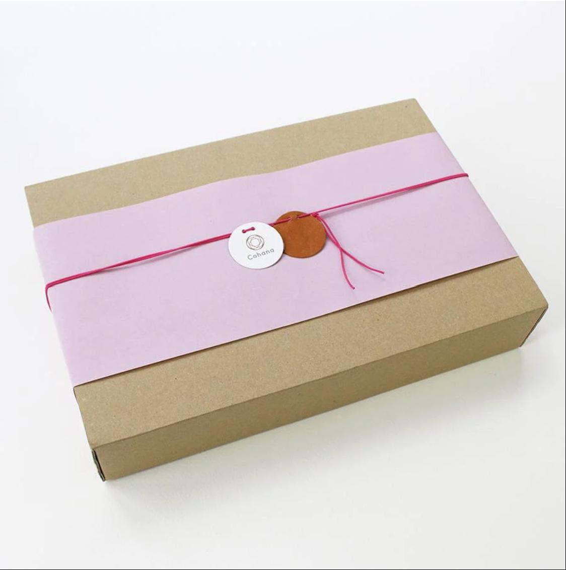 A brown cardboard box with a pink ribbon and a white circular tag with a logo on it, placed on a white background.