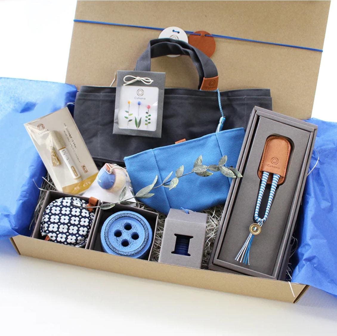 A gift box containing various sewing and crafting supplies, including a gray tote bag, a blue and white patterned bag, a blue button, a gray box with a blue and white pattern, and a gray box with a blue and white pattern.