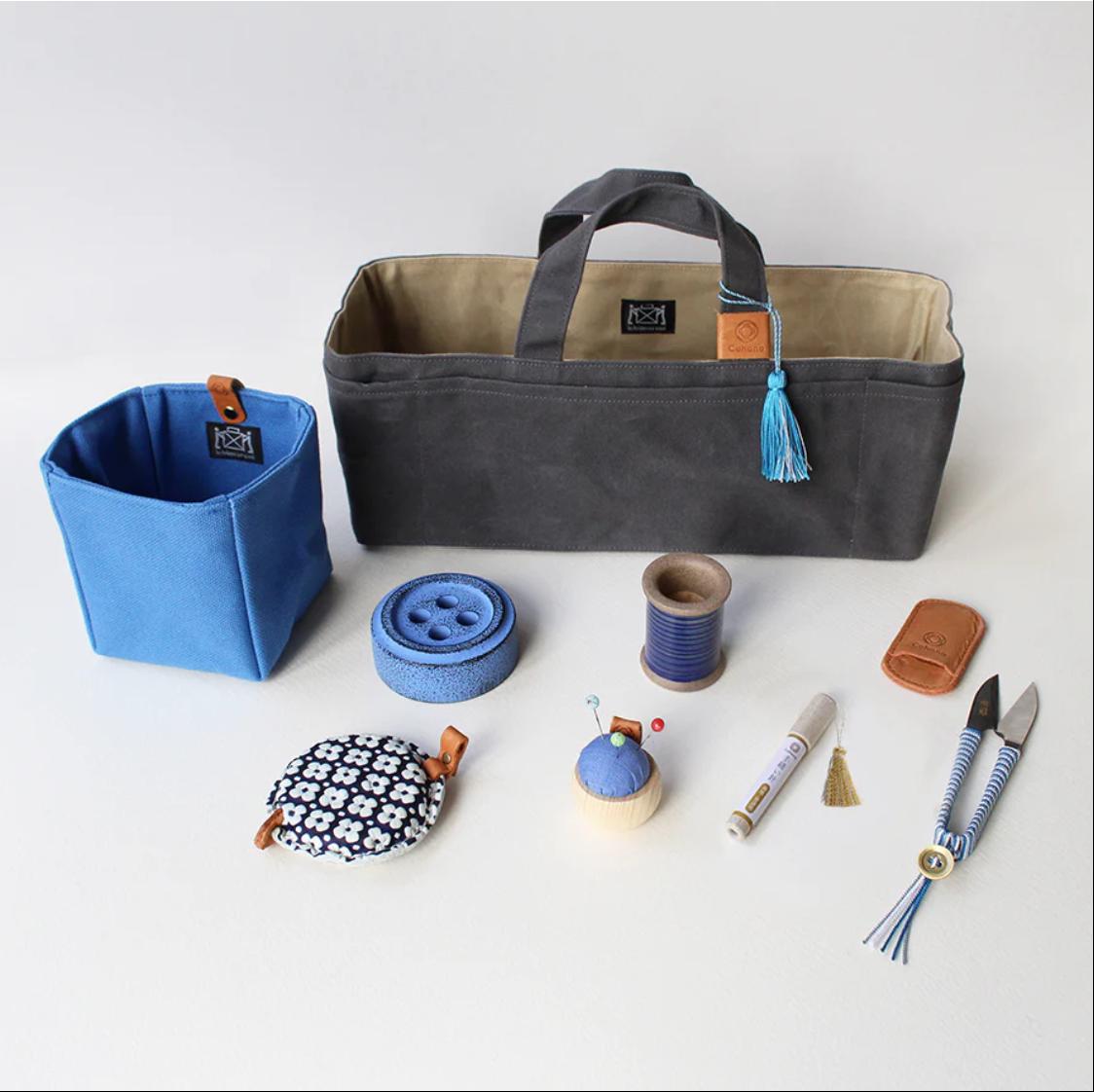 a collection of sewing and crafting supplies, including a gray tote bag, a blue container, a blue button, a blue spool of thread, a pair of scissors, a pair of pliers, a white marker, and a pair of scissors with a tassel.