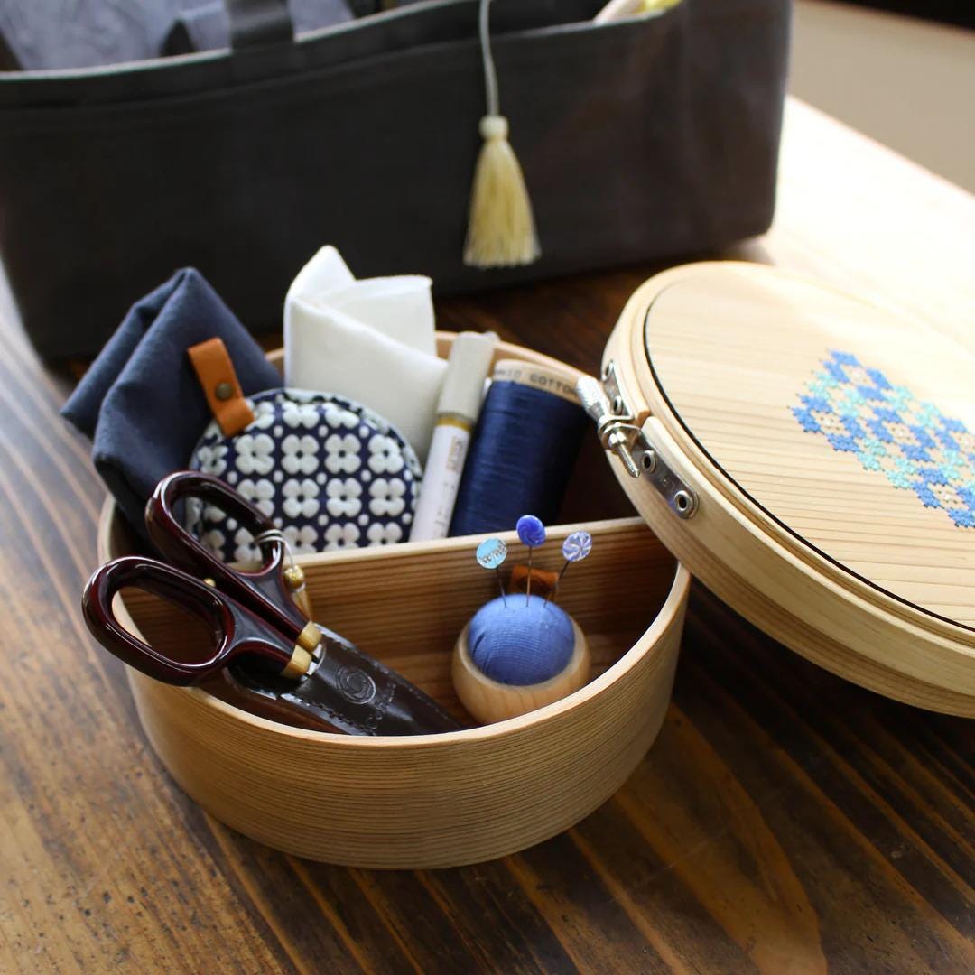A wooden container filled with sewing and crafting supplies, including a pair of scissors, a needle, thread, pins, and a tassel, sits on a wooden surface.