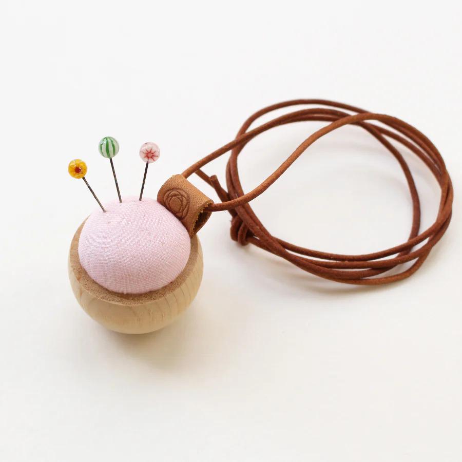A wooden pin cushion with colorful buttons on top, attached to a brown leather cord.