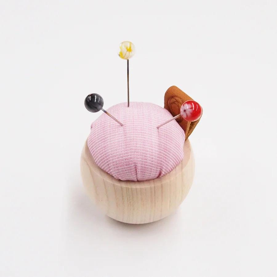 A pink and white checkered pin cushion with three colorful pins sticking out of it, placed on a white surface.