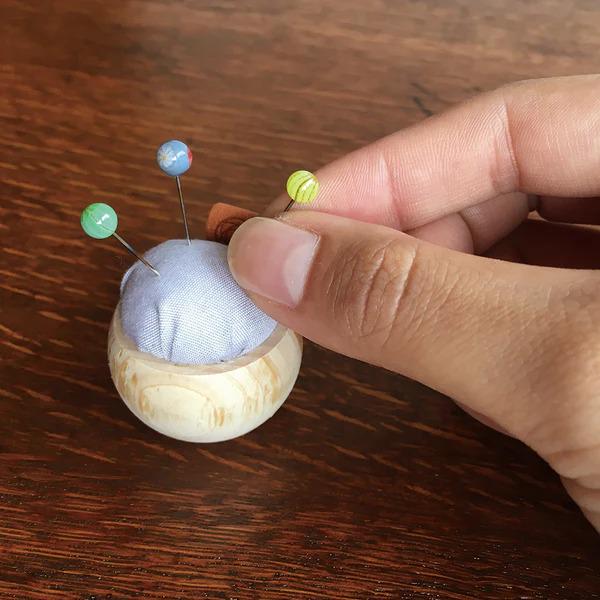 A hand is holding a small wooden pin cushion with colorful pins in it, placed on a wooden surface.